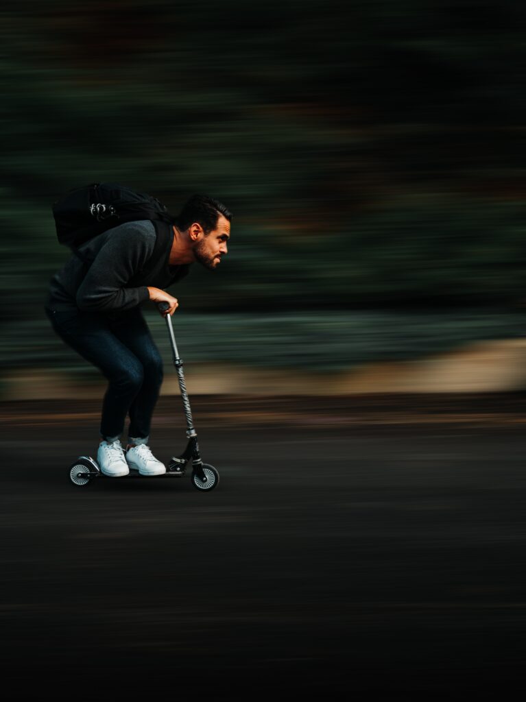 A person going fast on a scooter. The trees and bushes by their path are blurred because of the speed they're moving.
