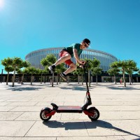 A person jumping over a scooter. Behind them is a circular building and clear blue skies.
