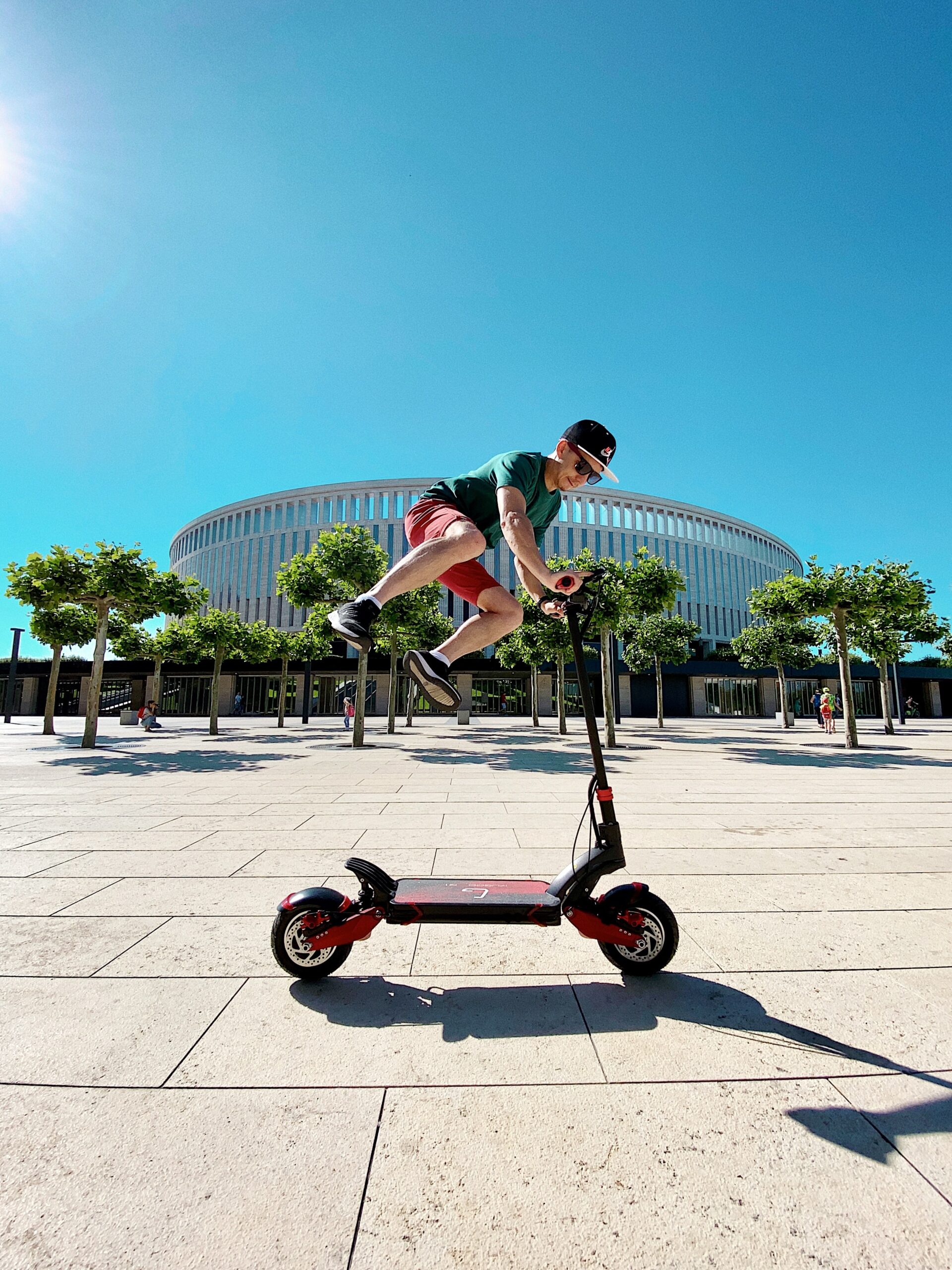 A person jumping over a scooter. Behind them is a circular building and clear blue skies.