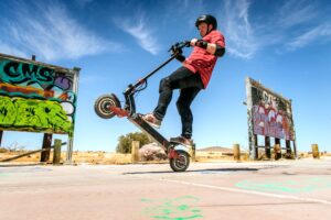 A person on a scooter doing a wheelie. Behind them is an empty field and mostly clear, blue skies.