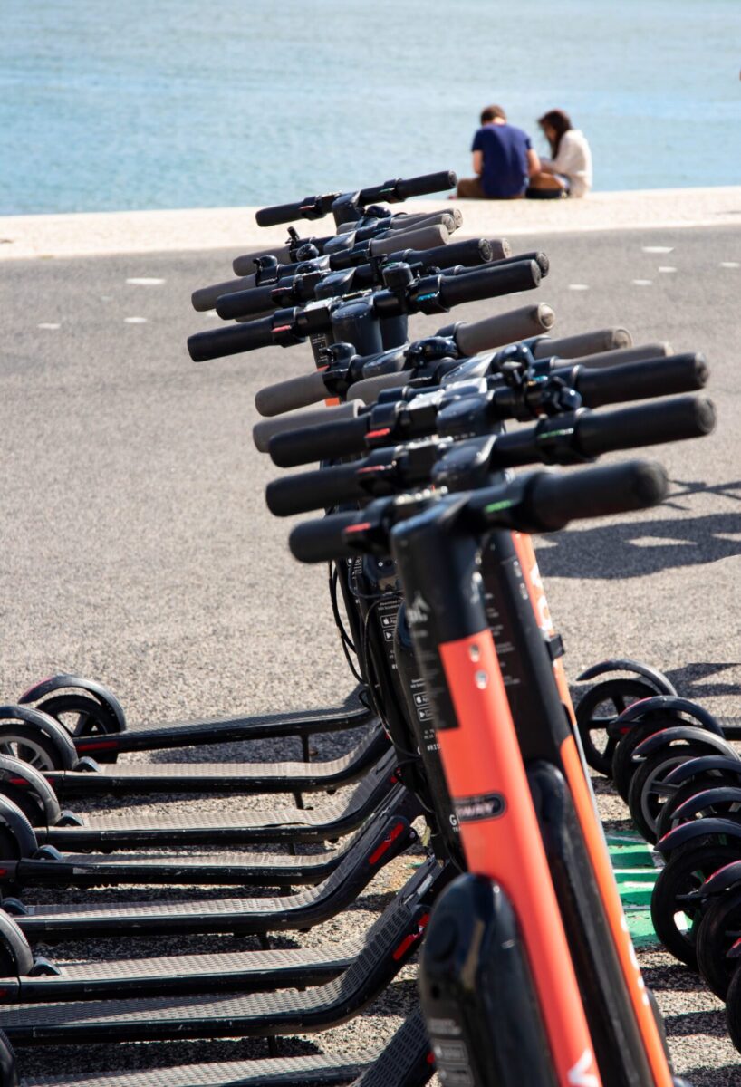 Row of parked scooters on the beach, in front of two people sat on the beach, looking at the waves in front of them.